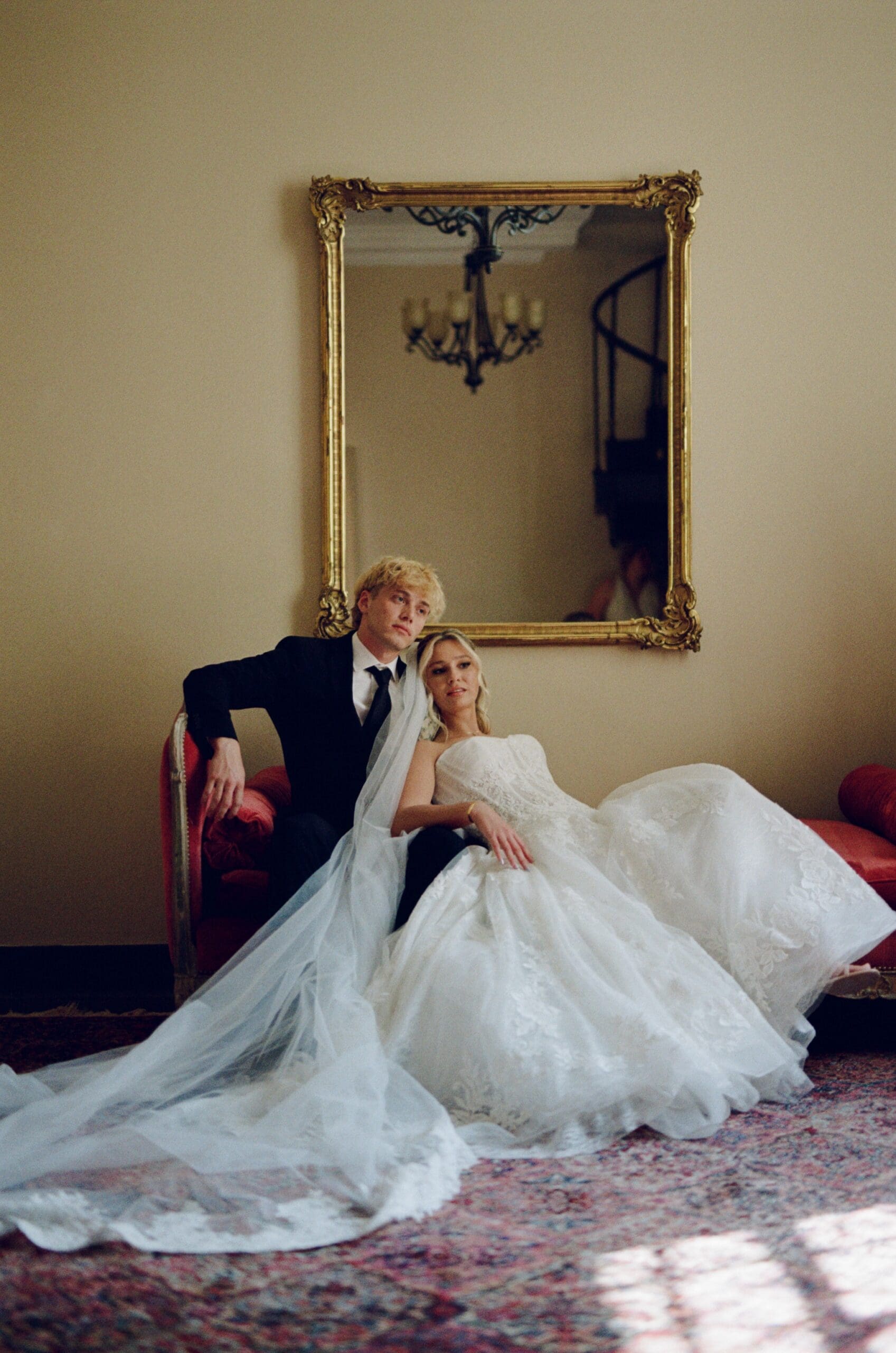 Bride and groom seated together on a vintage red settee, posing for an elegant indoor wedding portrait beneath an ornate gold mirror.