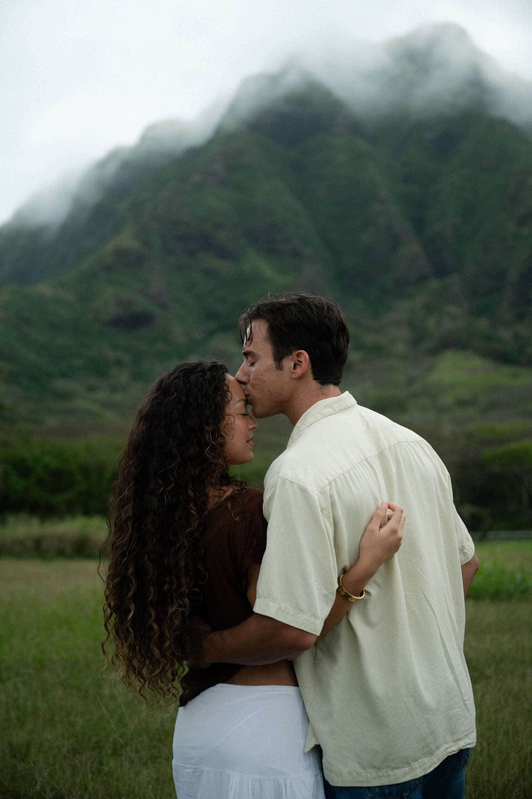 A couple stands at the base of a mountain and the man kisses his girlfriend on the forehead in Oahu, Hawaii