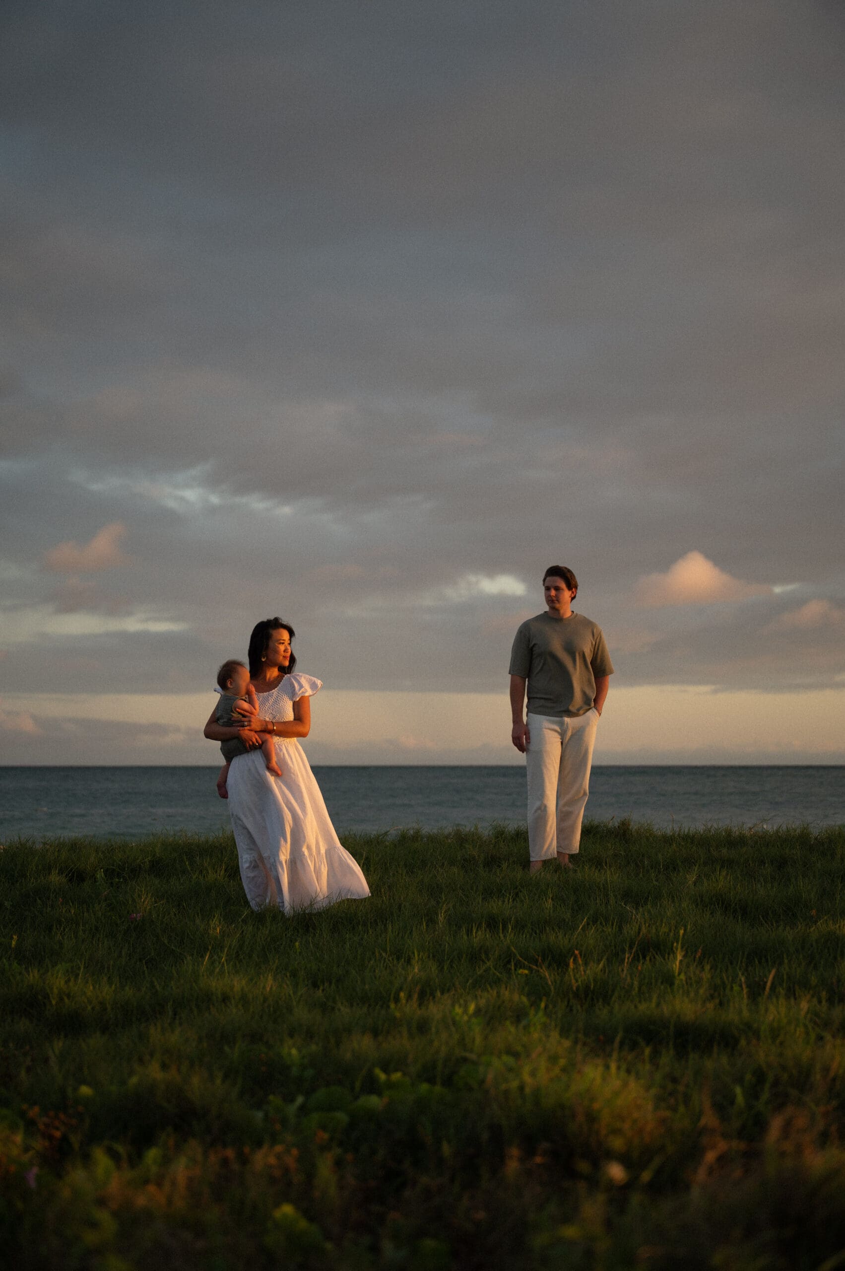 A husband, wife and baby stand on a grassy tuft with the ocean in the background at sunset