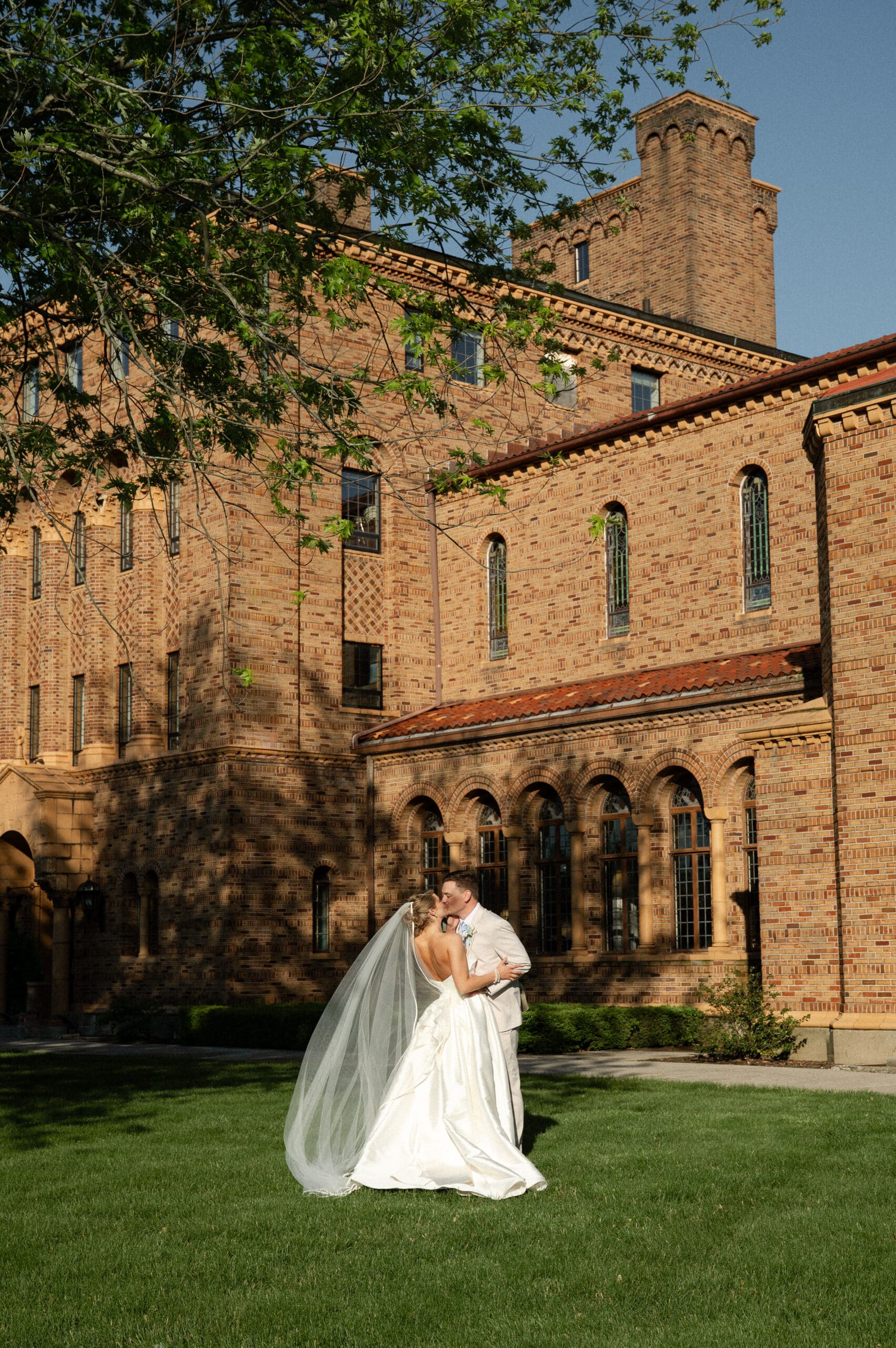 Bride and groom sharing a kiss on a lawn outside a historic brick estate during golden-hour wedding portraits.
